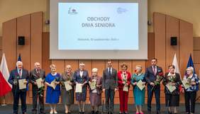 Pokaż zdjęcie: Na scenie stoi grupa seniorów, trzymających róże oraz dyplomy, obok nich znajdują się organizatorzy wydarzenia. W tle widoczny jest ekran z napisem "Obchody Dnia Seniora, Białystok, 02 października 2024 r.". Po bokach umieszczone są flagi Polski i Unii Europejskiej.