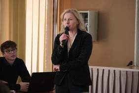 Pokaż zdjęcie: Zdjęcie kobiety, blondynki ubranej w czarną marynarkę, kobieta trzyma w ręku mikrofon.