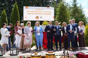 Pokaż zdjęcie: Uczestnicy uroczystości stoją na scenie i odbierają wyróżnienia; w tle baner jubileuszu PSONI, mikrofony i zielone otoczenie ogrodu.