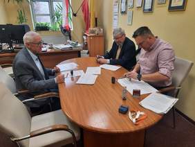 Pokaż zdjęcie: W gabinecie znajduje sie trzech mżęczyzn. Siedzą przy stole i podpisują umowę. Przed nimi leżą różne domumenty. Jeden z panów przystawia pieczątkę do umowy. 