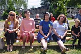 Pokaż zdjęcie: na trawie na długich ławkach siedzą kobiety, mężczyźni i dzieci