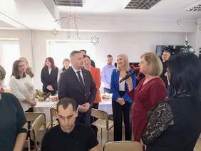 Pokaż zdjęcie: Spotkanie opłatkowe, pomiędzy stołami stoją goście, przy mikrofonie kobieta blondynka ubrana w bordową sukienkę.