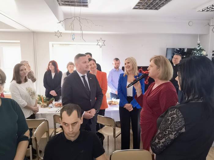 Pokaż zdjęcie: Spotkanie opłatkowe, pomiędzy stołami stoją goście, przy mikrofonie kobieta blondynka ubrana w bordową sukienkę.