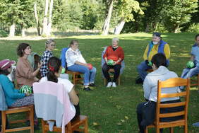 Pokaż zdjęcie: W słoneczny dzień, w parku na zielonej trawie, na drewnianych krzesłach ustawionych w okrąg siedzą osoby trzymające w rękach piłki terapeutyczne na wzór arbuza. Patrzą się w stronę kobiety, która ma podniesioną dłoń i mówi do nich. W tle 