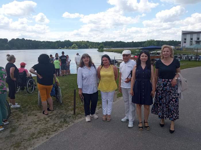 Pokaż zdjęcie: Przy jeziorze na chodniku stoi grupa pięciu osób w ty jeden mężczyzna, pozują do zdjęcia. W tle jezioro i inni ludzie