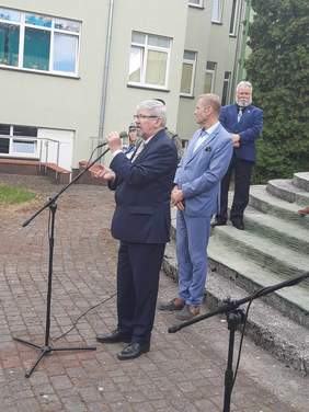 Pokaż zdjęcie: Na schodach stoi pan w garniturze, który wygłasza przemówienie do mikrofonu. Za nim stoją inni ludzie. W tle widać budynek. 