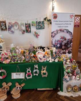 Pokaż zdjęcie: Na stole przykrytym zielonym obrusem leży mnóstwo ozdób i dekoracji wielkanocnych. Po prawej stronie stoi baner Polskiego Stowarzyszenia Ludzi Cierpiących na Padaczkę Oddział Łódzki w Ozorkowie. 