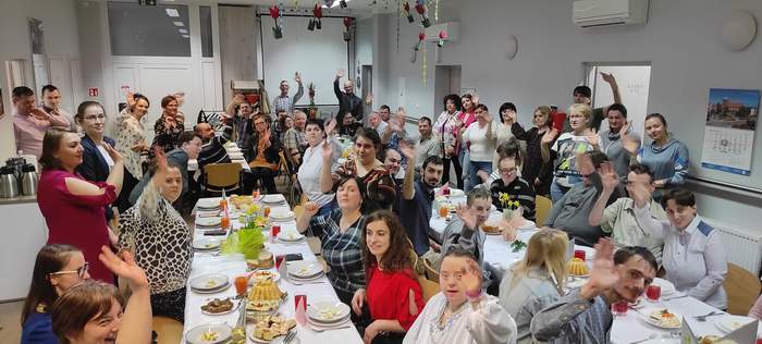 Pokaż zdjęcie: Zdjęcie przedstawiające uczestników WTZ przy stole z uniesionymi rękami w geście radości