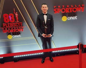 Pokaż zdjęcie: Szymon Sowiński na tle szarym, w garniturze, uśmiechnięty, trzyma w rękach statuetkę za nagrodę Sportowca Roku