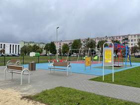 Pokaż zdjęcie: Na terenie miejskim znajduje się ogrodzony plac zabaw dla dzieci. Są na nim urządzenia, z których mogą korzystać także osoby z niepełnosprawnościami. W tle widać osiedle mieszkaniowe. 