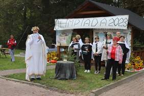 Pokaż zdjęcie: Zdjęcie na dworze osoba przebrana za Zeusa mówi do mikrofonu, z boku stoją zawodnicy.