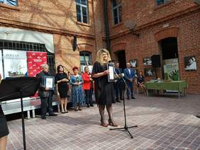 Kobieta w czarnej sukience, kręcone blond włosy stoi przed mikrofonem, za nią na tle budynku z cegły grupa ludzi