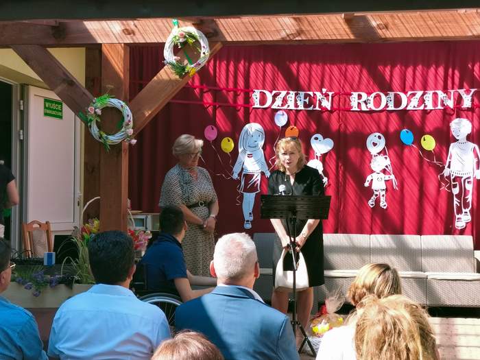 Pokaż zdjęcie: Na scenie przy mikrofonie stoi kobieta i przemawia, z tyłu za plecami na kurtynie jest napis piknik rodzinny