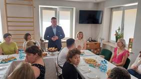 Pokaż zdjęcie: Mężczyzna w garniturze przemawia podczas zakończenia roku szkolnego, pozostałe osoby siedzą przy stole. 