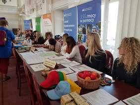 Ciąg stołów, za nimi przedstawiciele instytucji, za nimi banery, widoczne okna