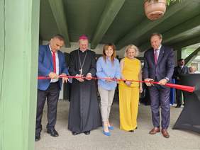 Pokaż zdjęcie: Pięć osób, w tym duchowny w czarnej sutannie z biskupią piuską, przecina czerwoną wstęgę podczas uroczystego otwarcia obiektu. Uczestnicy stoją pod zadaszeniem, trzymając nożyczki. Kobieta w żółtym stroju wyróżnia się na tle pozostałych osób w formalnych ubraniach.