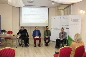 Pokaż zdjęcie: W sali konferencyjnej trwa panel dyskusyjny, na środku sali siedzi dwóch mężczyzn na wózkach i dwóch mężczyzn na krzesłach. Rozmawiają