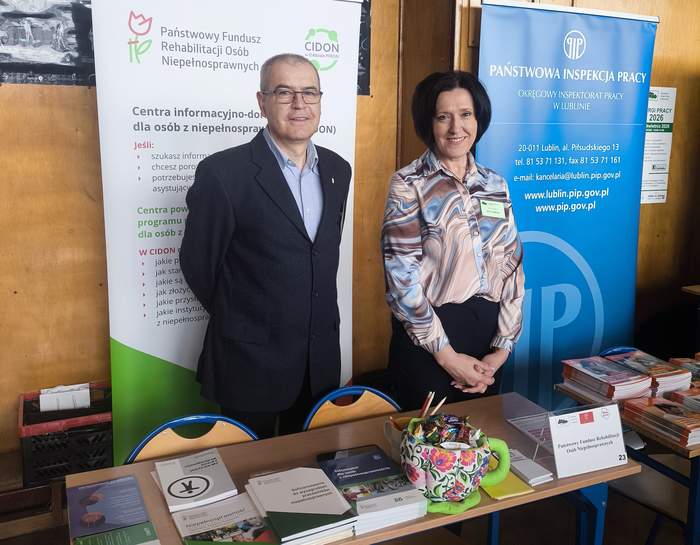 Pokaż zdjęcie: Na tle roll-upu stoi mężczyzna i kobieta. W tle widoczny roll-up PFRON i plakat Centrum Informacyjno-Doradczego.