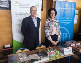Pokaż zdjęcie: Na tle roll-upu stoi mężczyzna i kobieta. W tle widoczny roll-up PFRON i plakat Centrum Informacyjno-Doradczego.