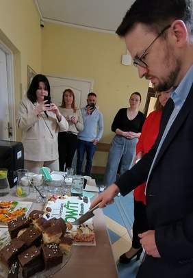 Pokaż zdjęcie: Mężczyzna w okularach i garniturze kroi tort z napisem „OWIT” na stole zastawionym słodkościami. W tle kilka osób obserwuje i fotografuje moment, a ściany pomieszczenia są pomalowane na żółto.