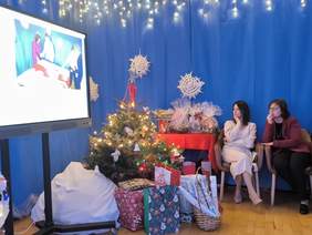 Pokaż zdjęcie: Pomieszczenie udekorowane w świątecznym klimacie. Po lewej stronie widoczny jest monitor z wyświetlonym zdjęciem, w centrum znajduje się ozdobiona choinka, a wokół niej ułożone są prezenty. Po prawej stronie na fotelach siedzą dwie osoby, obserwujące wydarzenie.