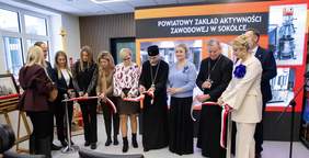 Pokaż zdjęcie: Grupa przedstawicieli władz i duchowieństwa przecina biało-czerwoną wstęgę podczas oficjalnego otwarcia Powiatowego Zakładu Aktywności Zawodowej w Sokółce; w tle widoczny ekran z nazwą placówki.