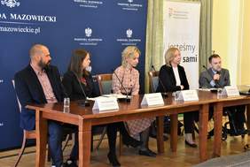 Spotkanie prasowe urzędu wojewódzkiego, osoby za stołem na tle ścianki z napisem wojewoda mazowiecki