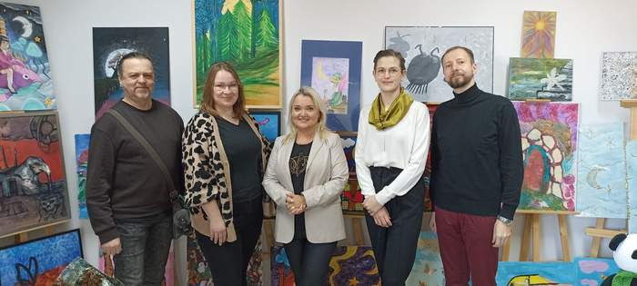 Pokaż zdjęcie: Kilka kobiet i mężczyzna stoją obok siebie na tle prac plastycznych konkursu "Sztuka Osób Niepełnosprawnych", które znajdują się w pomieszczeniu Oddziału PFRON w Zielonej Górze