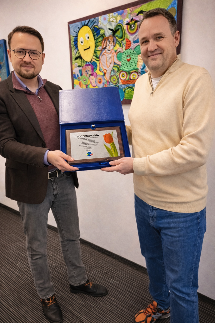 Pokaż zdjęcie: Dwóch uśmiechniętych mężczyzn stoi w pomieszczeniu na tle kolorowego obrazu, wspólnie prezentując otwarte, ozdobne etui z pamiątkowym dyplomem ozdobionym grafiką tulipana.