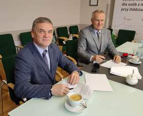 Pokaż zdjęcie: Dwóch mężczyzn elegancko ubranych siedzących przy stole. Na stole leżą dokumenty.