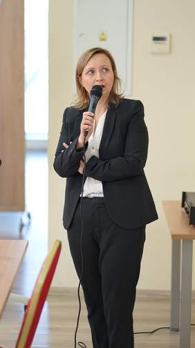 Pokaż zdjęcie: Zdjęcie podczas wygłaszanej prelekcji, kobieta blondynka ubrana w granatowy garnitur i białą bluzkę ma skrzyżowane ramiona, w ręku trzyma mikrofon.