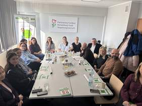 Pokaż zdjęcie: Na ekranie widać długi biały stół ustawiony w prostokąt, a wokół niego siedzi kilkanaście osób. Na stole stoją szklanki, butelki z wodą i napojami, kubki, materiały szkoleniowe oraz talerz z przekąskami na środku. W tle na ścianie wisi baner z napisem „Państwowy Fundusz Rehabilitacji Osób Niepełnosprawnych”. Po lewej stronie stoi dodatkowy baner z logo „OWiT Kalisz”. Po prawej stronie wisi kilka kurtek na wieszakach.