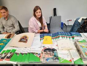 Pokaż zdjęcie: Młoda kobieta siedzi przy stoisku. Przed nią ustawiony jest stół z materiałami promocyjnymi i informatorami PFRON. Po lewej stronie widać siedzącego mężczyznę.  