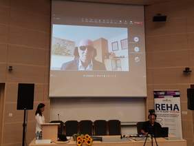 sala konferencyjna, przy stole siedzi mężczyzna, za nim ekran widać mężczyznę w ciemnych okularach, przemawia, z lewej strony za mównicą kobieta