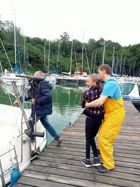 mężczyzna wchodzi na jacht, na podeście mężczyzna w żółtych spodniach podtrzymuje uśmiechniętą dziewczynę