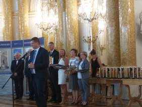 Pokaż zdjęcie: Mężczyzna stojący przy mikrofonie, w tle grupa osób trzymając w rękach statuetki