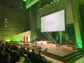 Pokaż zdjęcie: Na scenie stoi mężczyzna ubrany w garnitur. Trzyma list oprawiony w twardą okładkę i mówi do mikrofonu. Obok stoi kobieta w blond włosach i w czerwonej sukni, i mężczyzna ubrany w garnitur białą koszulę i muchę. Na telebimie wiszącym na scen