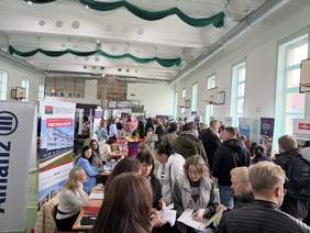 Pokaż zdjęcie: W hali sportowej liczne stoiska, uczestnicy rozmawiają i przeglądają materiały. Widoczne banery. Duże okna, kosze do koszykówki i tłum odwiedzających.