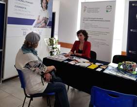 Pokaż zdjęcie: Przy stoliku na którym leżą ulotki i foldery siedzą dwie panie i rozmawiają. 