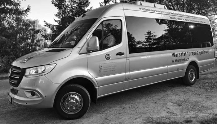 Pokaż zdjęcie: Na tle drzew iglastych widoczny jest autobus z naklejkami informującymi, że pojazd dofinansowany został ze środków PFRON i jest dostosowany do przewozu osób na wózku. Dodatkowo na autobusie możemy dostrzec dwa napisy: Warsztat Terapii Zajęciowej w Wiardunkach oraz Wielkopolskie Stowarzyszenie Na Rzecz Chorych, Niepełnosprawnych i ich Rodzin „Rehabilitacja” im. Karola Marcinkowskiego w Wągrowcu.