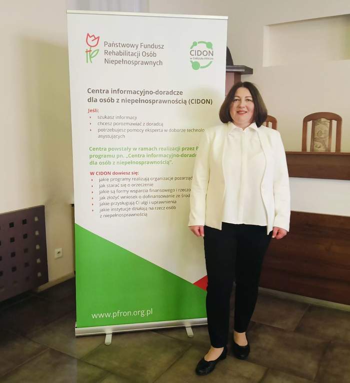 Pokaż zdjęcie: Na tle rollupu z logo CIDON i PFRON pozuje kobieta z ciemnymi półdługimi włosami w białej marynarce i ciemnych spodniach.