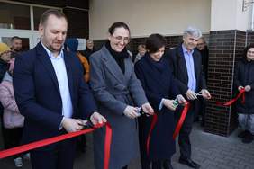Pokaż zdjęcie: Grupa czterech elegancko ubranych osób przecina czerwoną wstęgę. W tle, bezpośrednio przy wejściu do budynku, stoi większa grupa osób, wśród których znajdują się uczestnicy Środowiskowego Domu Samopomocy w Odolanowie.