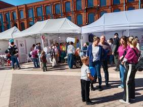 Pokaż zdjęcie: Na Rynku łódzkiej Manufaktury znajduje się tłum ludzi. Spacerują, jedzą lody, rozmawiają. Są dzieci i osoby z niepełnosprawnościami. 