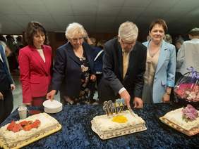 Pokaż zdjęcie: Na stole stoją trzy torty. Mężczyzna kroi jeden z nich. Obok pana stoją trzy kobiety, któe sie uśmiechają. W tle widać inne osoby. 