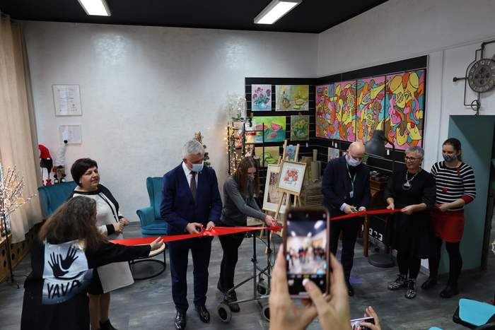 Pokaż zdjęcie: Fragment pomieszczenia trzy kobiety trzymają rozciągniętą czerwoną wstęgę, jedna kobieta się przygląda, a dwóch mężczyzn i dziewczyna trzymają w ręku nożyczki i przecinają wstęgę.