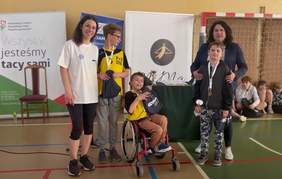 Pokaż zdjęcie: Na zdjęciu jest trze chłopców, jeden z nich siedzi na wózku. Wszyscy maja zawieszone medale. Obok nich stoją kobiety jedna ubrana na sportowo. Za nimi baner z Logo PFRON