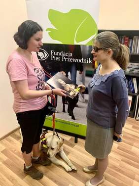 Pokaż zdjęcie: Na tle roll-up'u z napisem "Fundacja Pies Przewodnik" stoją dwie kobiety. Jedna z nich wręcza drugiej certyfikat. Pomiędzy nimi na podłodze leży pies rasy labrador w kolorze biszkopt.