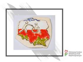 Pokaż zdjęcie: pocztówka z dziełem ubiegłorocznego laureata konkursu - płaskorzeźba z Polską w kolorach flagi (biało-czerwonym) i osobą na wózku 