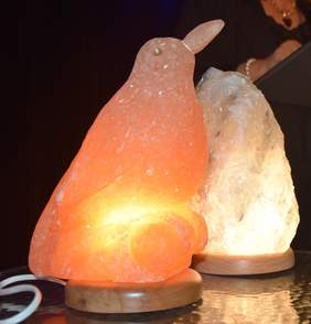 Pokaż zdjęcie: Na zdjęciu dwie figurki solne – jedna pomarańczowa w kształcie kosa, druga jasna w nieregularnej formie – stoją na stole z ciemnym blatem. Obie figurki mają podstawy z jasnego drewna i są podświetlone od środka