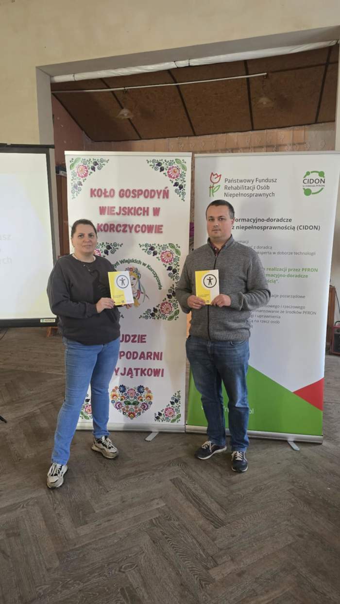 Pokaż zdjęcie: Kobieta i mężczyzna stoją obok siebie na tle banerów z logo instytucji i trzymają w rękach broszury o Dostępności